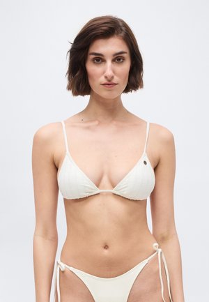 Donna con capelli castani corti che indossa un top bikini bianco testurizzato e un bikini con i lati annodati coordinato su uno sfondo uniforme.