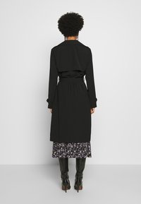 Trench-coat noir avec ceinture nouée, manches longues et détail à rabat dans le dos. Porté sur une jupe à motif floral et des bottes noires montantes jusqu'aux genoux.