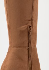 Brązowe zamszowe botki do kolan z bocznym zamkiem błyskawicznym, o gładkiej fakturze z widocznymi detalami ściegów wzdłuż szwu.