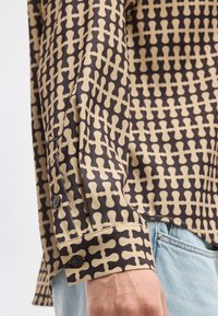 Μανίκι καφέ και μπεζ πουκαμίσου με σχέδια και μαύρα κουμπιά, σε συνδυασμό με ανοιχτόχρωμο μπλε τζιν.