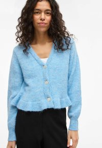 Hellblauer Cardigan aus weichem, strukturiertem Stoff. Mit V-Ausschnitt, vier Knöpfen und einem gerafften Saum sowie langen Ärmeln.