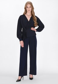 Blusa avvolgente blu navy con maniche lunghe, caratterizzata da un davanti annodato e una texture morbida, abbinata a pantaloni larghi coordinati e tacchi alti neri.