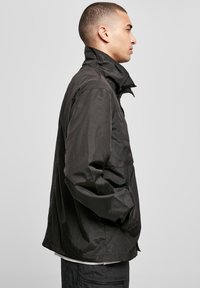 Juodas lengvas švarkas su aukštu kraštu ir ilgomis rankovėmis, turintis lygaus paviršiaus, priekinį užtrauktuką ir šonines kišenes.