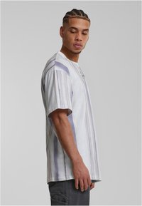 Braidelt hajú férfi fehér és szürke csíkos pólót és sötét nadrágot visel, háttérben egy sima szürke fal áll.