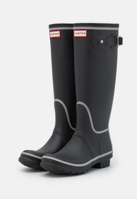 Svarta gummistövlar med rund tå, grå kant och spänne. Texturerade sulor för grepp och två logotyper på framsidan.