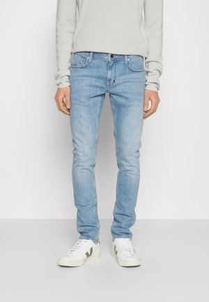 Person trägt hellblaue Slim-Fit-Jeans, weißes Langarmhemd und weiße Sneakers vor einem schlichten Hintergrund.