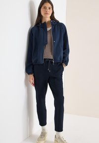 Veste bleu marine avec fermeture éclair frontale, ourlet élastique et capuche avec cordon, assortie à un pantalon assorti et des baskets de couleur claire.