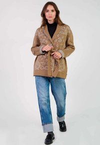 Gilet en tricot marron avec des motifs blancs abstraits, à l'avant ouvert, fermeture par lien, et poignets gris côtelés, associé à un jean bleu retroussé et des chaussures noires.