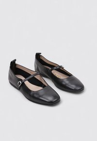 Bailarinas de charol negro con una sola tira delgada y hebilla plateada sobre una superficie gris claro.