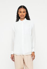 Woman with curly hair wearing a white button-up shirt with chest pocket and beige loose pants, standing against a plain white background.