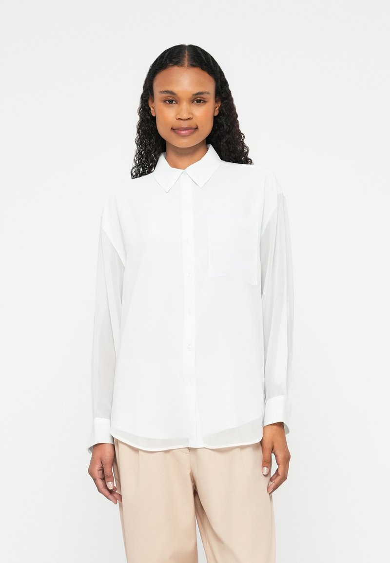 Woman with curly hair wearing a white button-up shirt with chest pocket and beige loose pants, standing against a plain white background.