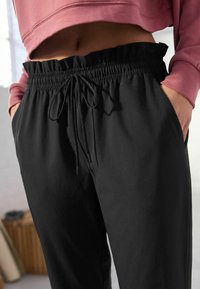 Pantalon noir en tissu lisse avec une ceinture élastique, une fermeture à cordon et des poches latérales. Le design présente une coupe décontractée.