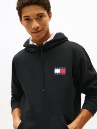 Junger Mann trägt einen schwarzen Hoodie mit einem "Tommy Jeans" Logo, darunter ein weißes Shirt, Hände in der Vordertasche, neutraler Hintergrund.