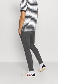 Homme portant une chemise rayée blanche et noire, un pantalon gris foncé, des chaussettes blanches et des baskets blanches avec des accents bordeaux et noirs, se tenant de côté.