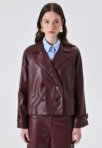 Vrouw in een bordeauxrood dubbelzijdig leren jack en bijpassende broek, lichtblauwe geknoopte blouse en grote gouden drop oorbellen, vooraanzicht.