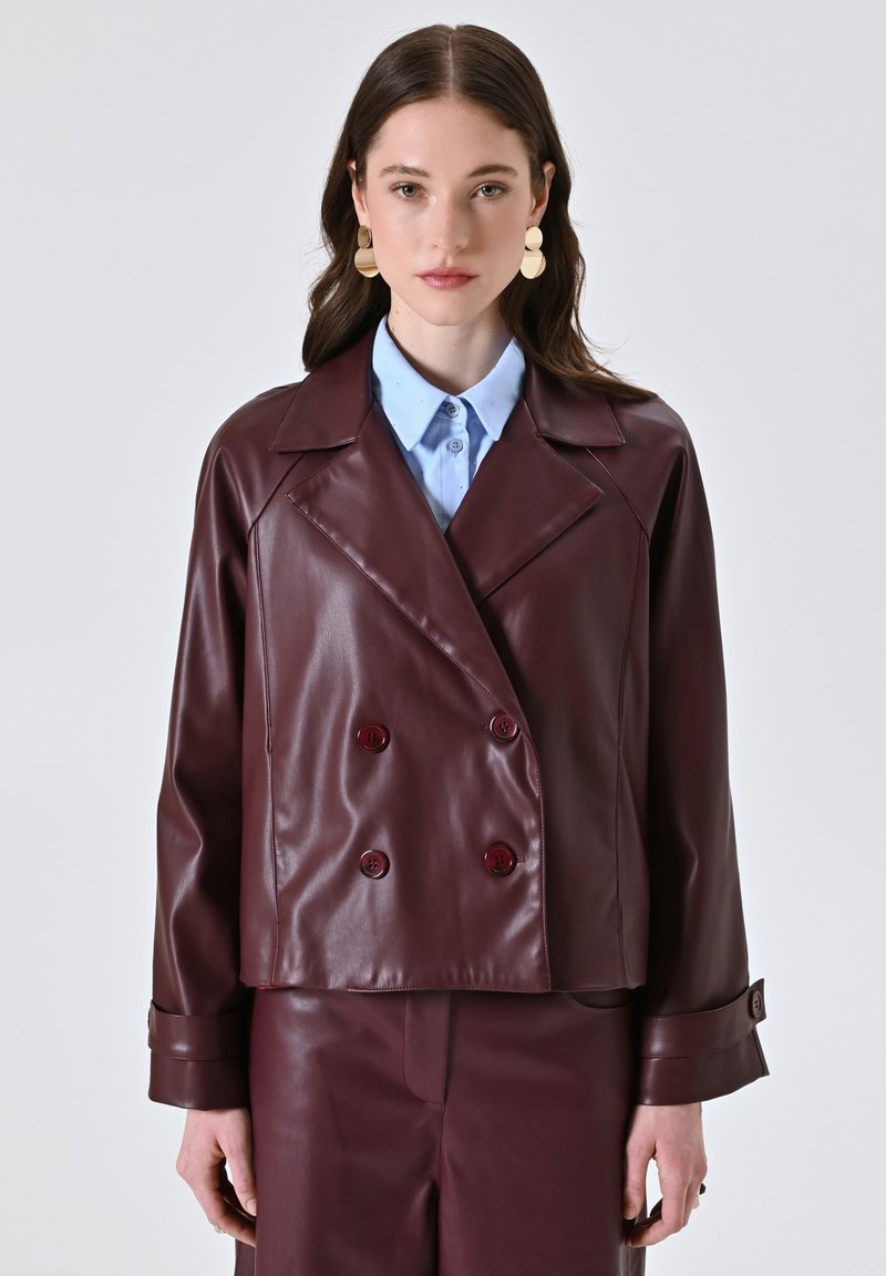 Vrouw in een bordeauxrood dubbelzijdig leren jack en bijpassende broek, lichtblauwe geknoopte blouse en grote gouden drop oorbellen, vooraanzicht.