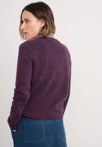 Pleteni pulover u tamnopurple boji s dušičastim šarenim mrljama, uskim rukavima i rebrastim rubom; nosi se preko plave rebraste suknje.