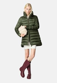 Grüne Steppdaunenjacke mit hohem Kragen, horizontalem Steppmuster und Reißverschluss. Modell hält cremefarbenes Pelzaccessoire und trägt burgunderrote Stiefel.