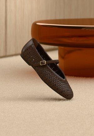 Σκούρο καφέ πλεκτό παπούτσι Mary Jane με χρυσή πόρπη, τοποθετημένο σε μπεζ χαλί δίπλα σε στρογγυλό τραπεζάκι χρώματος κεχριμπαριού.