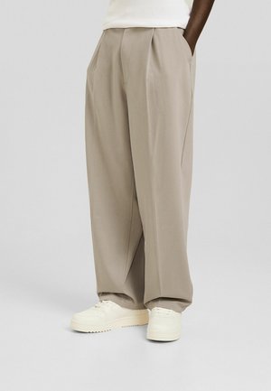 Persoon draagt losse beige broek en witte sneakers, staand met handen in de zakken tegen een lichtgrijze achtergrond.