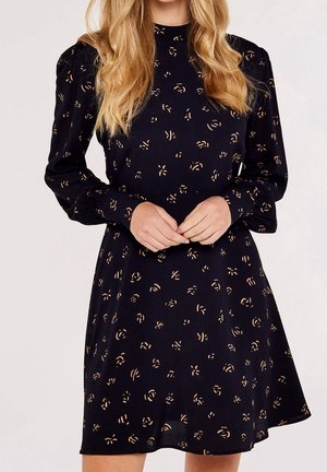 Kobieta o długich blond włosach, ubrana w czarną sukienkę z długimi bufkami na rękawach oraz małymi jasnobrązowymi abstrakcyjnymi wzorami kwiatowymi.