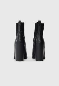 Black leather ankle boots with a thick, high heel and ribbed knit upper. Features a pull tab for easy wear. Smooth texture and sleek design.