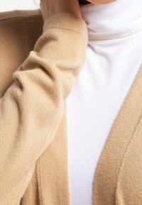 Cardigan beige en tissu doux, présentant une finition légèrement texturée, un design ouvert sur le devant et des manches longues superposées à un haut blanc.