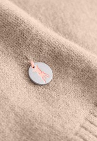 Ronde metalen tag met een grijze afwerking, met een roze silhouette van een koe. Geplaatst op een zachte, gestructureerde beige stof.