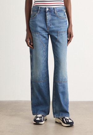 Personne portant un jean bleu patchwork à jambes larges avec poches avant, associé à des baskets noires et blanches et un haut rayé.