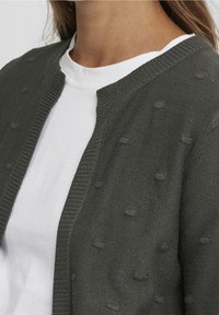 Szary dziergany kardigan z fakturowanym ściegiem bobble i prążkowanymi detalami wzdłuż dekoltu, noszony na zwykłej białej koszuli.