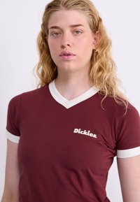 Burgunder T-Shirt mit weißem Abschluss am Ausschnitt und den Ärmeln. Verfügt über ein Logo im linken Brustbereich. Der Stoff scheint weich und figurbetont zu sein.