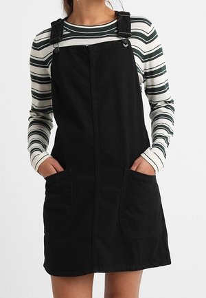 Vrouw draagt een zwarte tuinbroekjurk met twee voorzakken over een wit shirt met lange mouwen en zwarte en groene horizontale strepen.