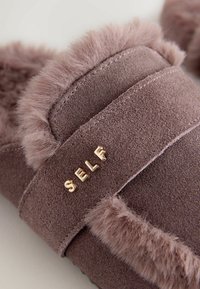 Lila Wildlederschuhe mit einem flauschigen Fellbesatz und einem goldmetallischen "SELF"-Detail an einem Riemen. Weicher Stoff und runde Form.