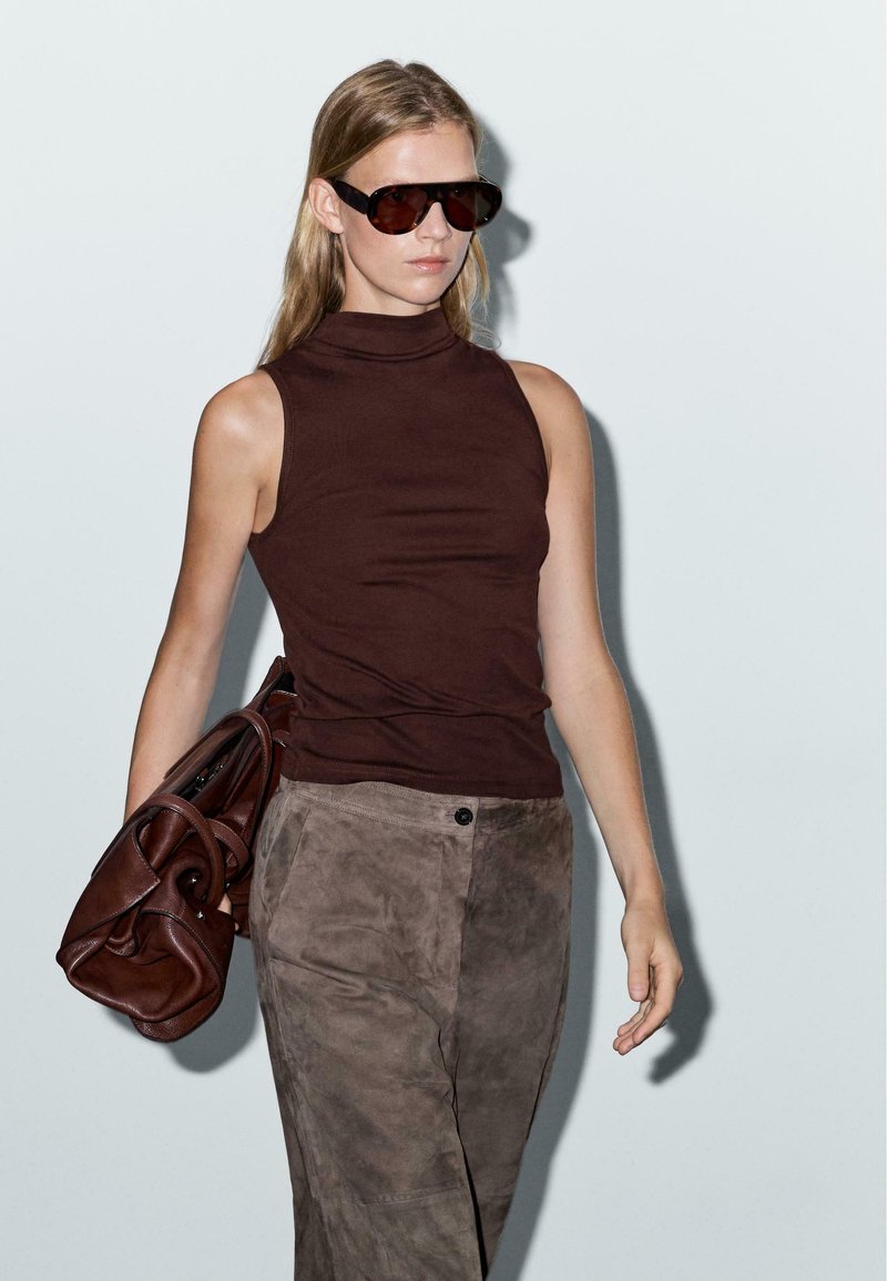 Haut sans manches marron, pantalon en suède vert olive, et grandes lunettes de soleil sombres. Le mannequin tient un sac à main marron, sur un fond clair.