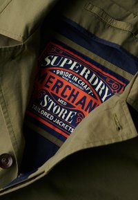 Veste ajustée olive avec une texture lisse, dotée d'une étiquette intérieure contrastante bleue et rouge qui indique "Superdry Merchant Store."