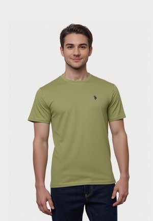 Junger Mann mit kurzen braunen Haaren, der ein olivgrünes kurzärmeliges T-Shirt und dunkelblaue Jeans trägt, vor weißem Hintergrund stehend.