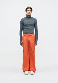 Grijze lange mouw zip-up top met oranje en blauwe accenten, gecombineerd met losse oranje cargo broek met zijzakken. Lichtgekleurde schoenen.