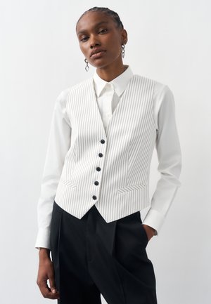 Μοντέλο φορεί γιλέκο λευκό με λεπτές ρίγες και μαύρα κουμπιά πάνω από λευκό πουκάμισο με γιακά και μαύρο παντελόνι, ποζάροντας με το ένα χέρι στην τσέπη.