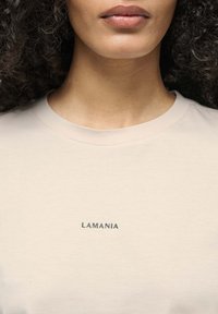 Světle béžové tričko z hladkého materiálu, s černým potiskem loga "LAMANIA" přes hruď. Jednoduchý, kulatý výstřih a krátké rukávy.