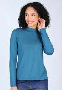 Tee in Teal mit langen Ärmeln und einem Stehkragen. Enganliegendes Design, gerippte Textur und eine glatte Oberfläche. Kombiniert mit Jeans.