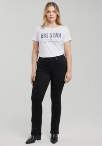 Žena se světlými vlasy, oblečená v bílém tričku s nápisem "BIG STAR", černých džínech s vysokým pasem a černých kotníkových botách, stojící před jednobarevným pozadím.