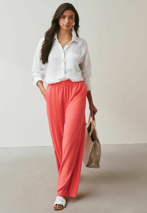 Hosszú sötét hajú nő fehér gombos ingben, korall színű bő szárú nadrágban, fehér szandálban, csíkos bevásárlótáskát hordva sétál.