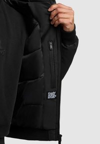 Jachetă neagră, căptușită, cu o textură netedă și mansete cu nervuri. Prezintă o buzunare interioare cu fermoar și o etichetă care spune "Khujo Original Changing Threads."