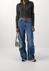 Sort cropped zip-up jakke, blå cargo jeans med lommer, sølv håndtaske og sort-hvide sneakers. Glatte teksturer og kontrasterende farver.
