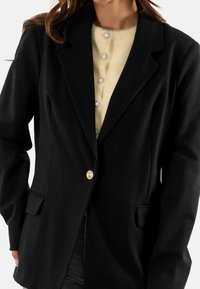 Blazer nero con un design classico, caratterizzato da rever a lancia, tasche frontali e un bottone dorato. Indossato sopra un top cremoso con texture e bottoni decorativi.