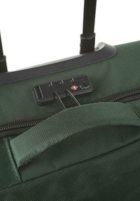 La bolsa de tela verde con ruedas presenta un candado combinado, cremalleras negras y un asa resistente. La textura está tejida con un patrón diagonal.