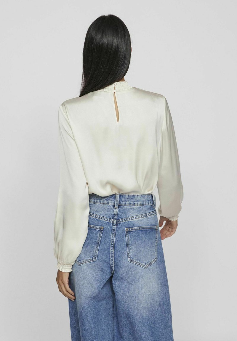 Blouse en satin crème avec un motif en keyhole au dos, associée à un jean en denim bleu clair taille haute avec des poches arrière.