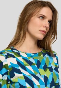 Junge Frau mit langem hellbraunem Haar, die ein mehrfarbiges, abstrakt gemustertes Oberteil in Blau-, Grün- und Weißtönen trägt und nachdenklich zur Seite blickt.