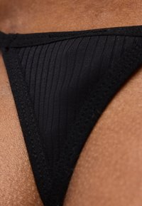 Gros plan sur un bas de bikini noir côtelé sur une peau foncée, montrant les coutures texturées et la forme triangulaire.