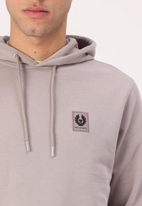 Γκρι φούτερ από απαλό ύφασμα, με κεντρική τσέπη καγκουρό, ρυθμιζόμενο κορδόνι και μαύρη ετικέτα λογότυπου στο στήθος.